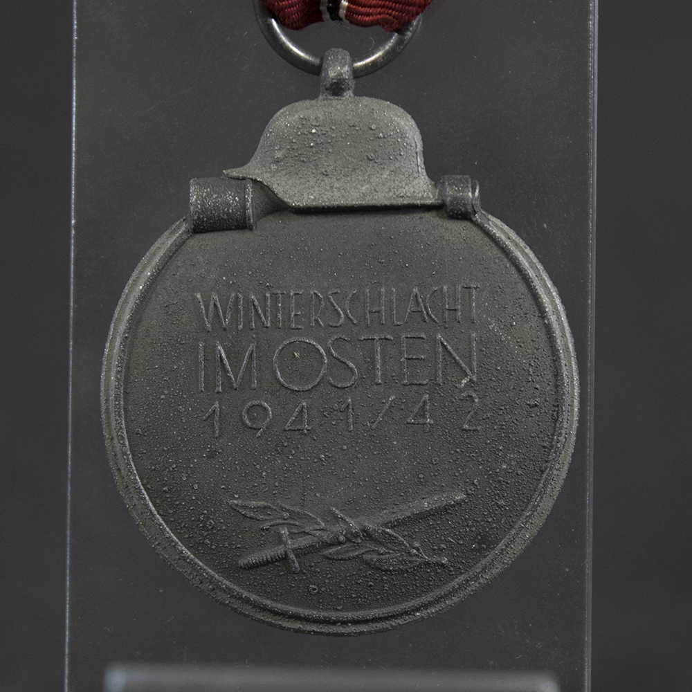 Close-up of the reverse of a WWII German Eastern Front Medal. The back displays the commemorative inscription and date in raised lettering, surrounded by a textured field. The surface exhibits original patina, light wear, and age-related toning.
