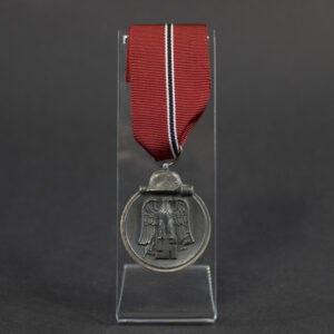 Full view of a WWII German Eastern Front Medal (Ostmedaille) suspended from its original red, black, and white ribbon. The medal displays the characteristic helmet-and-grenade design and shows age-related wear, dark patina, and minor edge wear. The suspension ring is maker-marked “93,” identifying the manufacturer as Richard Simm & Söhne.