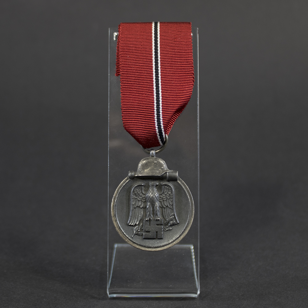 Full view of a WWII German Eastern Front Medal (Ostmedaille) suspended from its original red, black, and white ribbon. The medal displays the characteristic helmet-and-grenade design and shows age-related wear, dark patina, and minor edge wear. The suspension ring is maker-marked “93,” identifying the manufacturer as Richard Simm & Söhne.