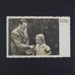 Black-and-white German photo postcard showing a public event scene with Adolf Hitler shaking hands with a young child. Several uniformed figures stand in the background. The upper right corner contains the German caption “Das Führers gütige Hand.” The postcard has a decorative scalloped border.