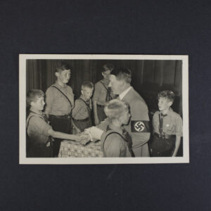 Black-and-white Photo-Hoffmann postcard showing Adolf Hitler shaking hands with a member of the Hitler Youth during an indoor gathering. Several Hitler Youth members in uniform surround a table while observing the presentation. The image is bordered by a white frame and printed as a real photographic postcard.