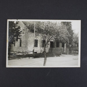 Black-and-white historical postcard depicting a residential house in Leonding, Austria. The building is shown from the street, surrounded by trees and a wooden fence. The photograph captures the exterior of the house identified as the parents' home in Leonding, with a tree positioned in the foreground.