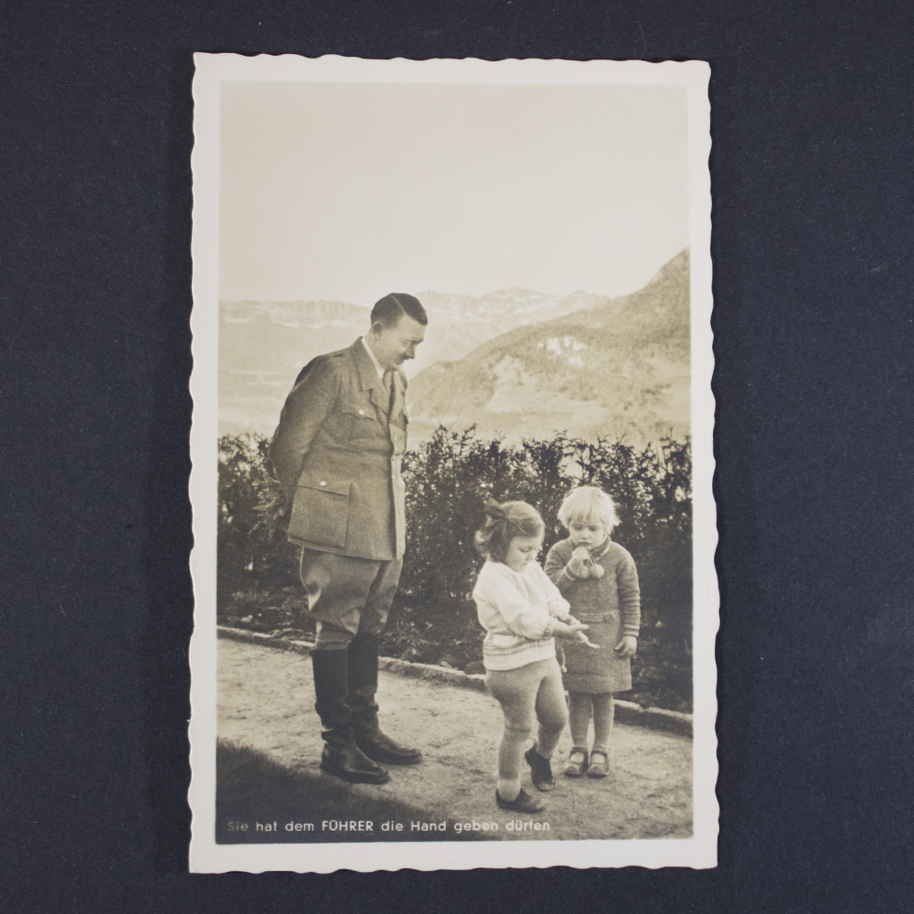 Black-and-white Photo-Hoffmann postcard showing a uniformed figure standing outdoors beside two young children on a garden path. Hills and mountains appear in the background. The postcard bears the German caption “Sie hat dem Führer die Hand geben dürfen” along the bottom edge and features a decorative scalloped border.