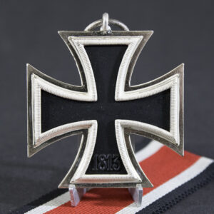 Reverse side of a World War II German Iron Cross 2nd Class medal featuring a plain black iron core with silver frame and the date “1813” on the lower arm. The medal is shown standing upright on its original black, white, and red ribbon.
