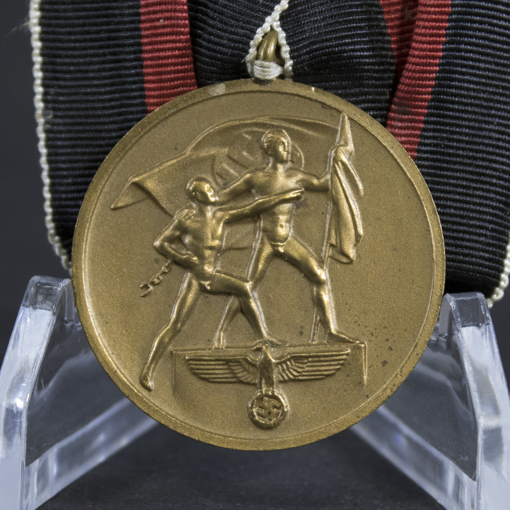 Close-up of a WWII German Sudetenland Medal showing two figures holding a flag, symbolizing the annexation of the Sudetenland. The bronze medal displays detailed relief artwork and is attached to its original folded ribbon medal bar.