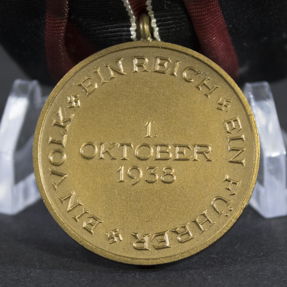 Close-up reverse view of a WWII German Sudetenland Medal showing the inscription “Ein Volk, Ein Reich, Ein Führer” around the edge and the date “1. Oktober 1938” in the center. The bronze medal is suspended from its original black, red, and white ribbon and displayed on a clear stand.