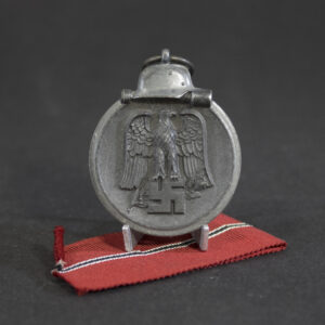 WWII German Eastern Front Medal (Ostmedaille) displayed upright on a stand with its original red, black, and white ribbon. The obverse features a Wehrmacht-style steel helmet above a national eagle clutching a swastika, surrounded by a circular border. The medal is maker marked “4” on the suspension ring.