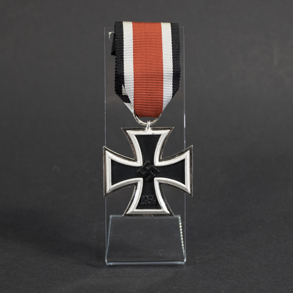 Close-up front view of a German Iron Cross 2nd Class (Eisernes Kreuz 2. Klasse) 1939 medal. The cross has a black center with a silver-colored border and features a raised swastika at the center. The year “1939” is visible on the lower arm. The medal is suspended from a metal ring and displayed on a clear acrylic stand against a dark background.