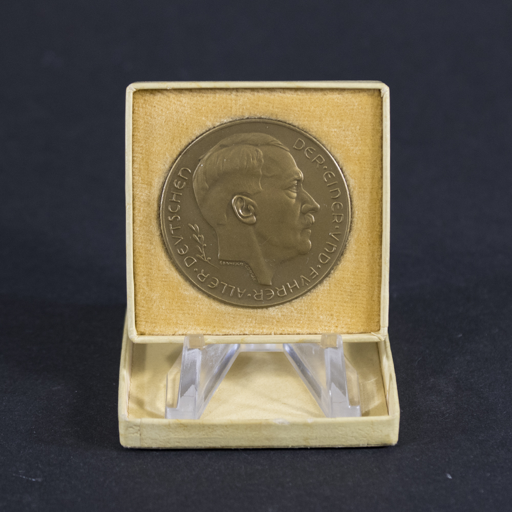 German Third Reich commemorative medal displayed inside an open cream-colored presentation case with a velvet-lined interior. The obverse side of the medal, featuring a right-facing portrait of Adolf Hitler and the inscription “DER EINER UND FÜHRER ALLER DEUTSCHEN,” is mounted on a clear acrylic stand against a dark background. The case shows light age-related wear and provides an attractive display for the medal.