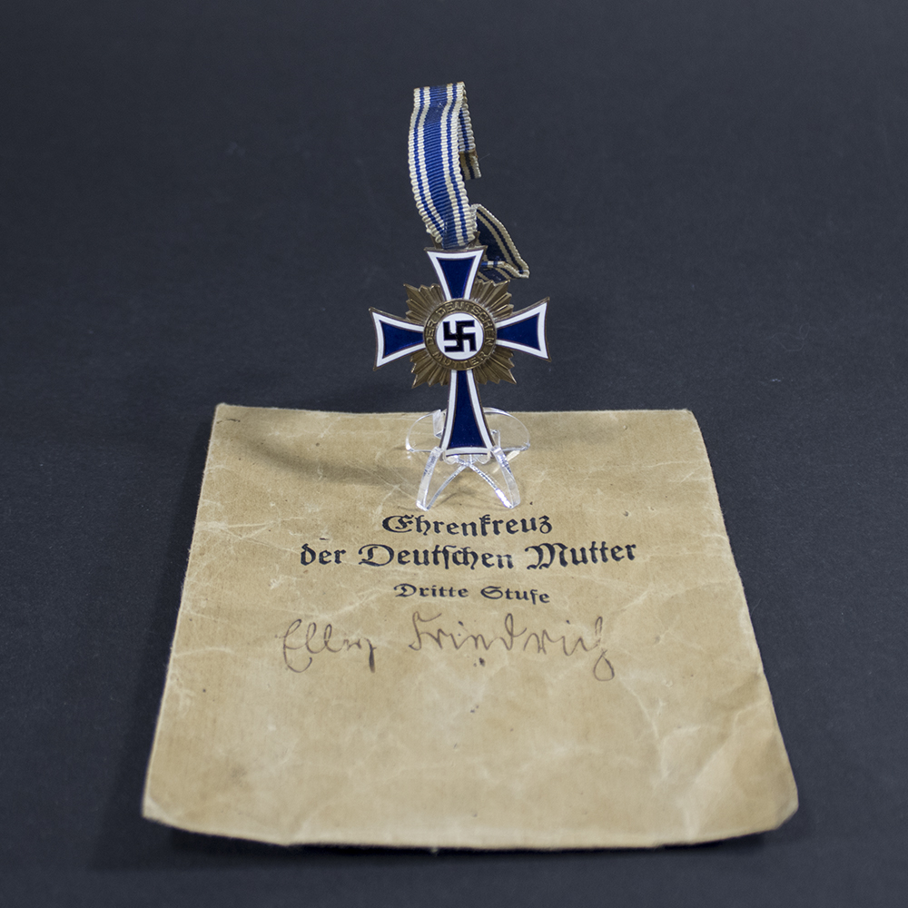 WW2 German Bronze Mother's Cross (Mutterkreuz 3rd Class) with blue and white ribbon displayed above its original paper issue envelope.